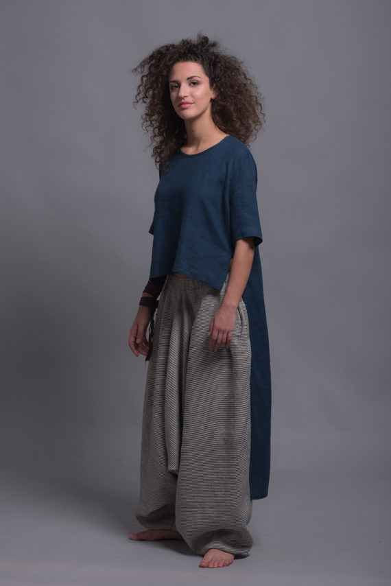 Model wearing the FRIDA linen shirt, emphasizing the shorter front and elongated back hemline with loose-fitting striped harem trousers, highlighting the wide leg and comfortable design.