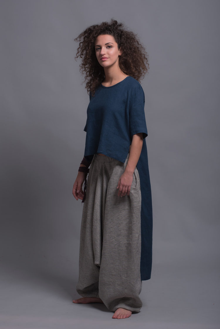 Model wearing the FRIDA linen shirt, emphasizing the shorter front and elongated back hemline with loose-fitting striped harem trousers, highlighting the wide leg and comfortable design.