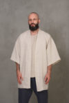 A front view of a man wearing a beige linen kimono-style jacket with short sleeves and an open front, styled over a casual outfit.